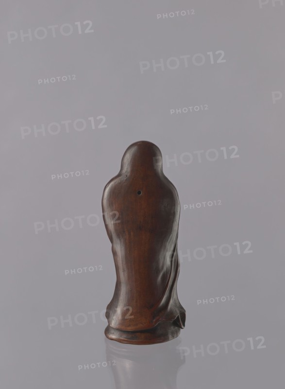 Daruma, Japanese, Meiji period 1868–1912. Ceramic figurine depicting Daruma Bodhidharma as a male Buddhist monk wearing headgear and a long flowing robe covering the head. Late 19th century, Seto, Japan. Height: 21.2 cm., A smooth, rounded sculpture viewed from the back, made of a polished dark brown material with subtle lighter brown variations and a small circular depression near the upper center; the form tapers downward into flowing, draped contours and rests on a slightly flared base, with a soft reflection of the piece visible on a pale gray surface against a uniform light gray background.