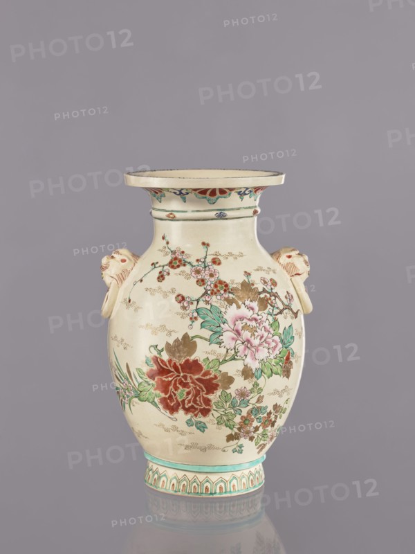 Vase, decorated with flowers and lions in satsuma ware; Japanese ceramics with cracklework decoration depicting peonies, chrysanthemums, and plum trees. Meiji period ca. 1870–1880. Vessel: height 26.5 cm; width 17.5 cm; diameter 16.5 cm., An off-white ceramic vase with a flared rim and a rounded body sits against a gray background; the vase features painted floral decorations in shades of red, pink, green, brown, and teal, with a large red flower and a pale pink flower amid green leaves and smaller blossoms, and delicate gold dotted accents scattered across the surface; there are sculpted handles on either side shaped like stylized animal heads in the same off-white with light brown shading, a decorative band with teal and brown motifs around the neck, and a patterned teal and brown border at the base, with the vase casting a faint reflection on the surface beneath it.