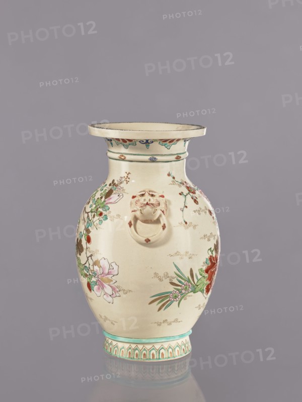 Vase, satsuma earthenware, decorated with crackle glaze and painted floral motifs including peonies, chrysanthemums, and plum trees, also featuring lions; Meiji period ca. 1870–1880, Japan; vessel; height 26.5 cm, width 17.5 cm, diameter 16.5 cm., An off-white ceramic vase with a flared rim and a rounded body sits against a muted gray background; the vase is decorated with hand-painted floral motifs in pink, red, green, and teal, with additional small brown and gold accent dots and fine brushwork; a sculpted lion-head ring handle in a similar off-white tone is attached to the upper body; the neck and base feature patterned bands in teal and beige with repeating geometric and foliate shapes; the vase casts a soft reflection on the surface beneath it.