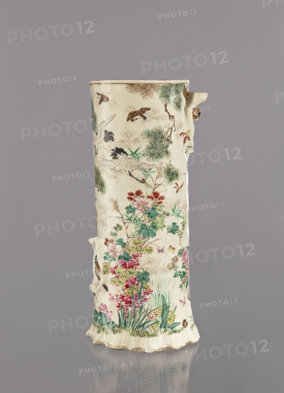 Vase, Tôritei; satsuma; Japanese; decorated with insects, tree trunks, flowers, plant ornaments, birds, peonies, pines, crickets, monkeys, kacho flowers and birds, kachôga iconography, lotuses, irises, grasses, cranes; Meiji period 1868–1912, ca. 1885 Meiji 18; Japan, Kyoto, Awata; vessel; height 32 cm, width 15 cm., A tall cylindrical ceramic vase with an uneven rim and textured surface, decorated with painted natural scenes; the body shows a group of birds in flight and perched among pine-like branches, and a dense cluster of flowering plants and grasses at the lower portion, including blossoms and leafy stems. The painted colors visible include cream as the base color, greens for foliage, reds and pinks for flowers, yellow and purple accents in blossoms, brown and tan tones in branches and some birds, white on some bird plumage, and small touches of black for detailing. The vase has a slightly irregular, handmade appearance with a few raised protrusions on the sides and a scalloped ...