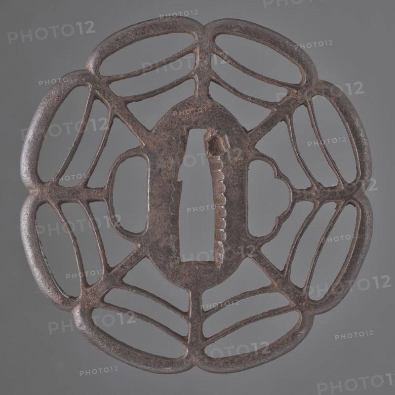 Crossguard tsuba, Tomotsugu; Japanese, Edo period 1603–1868, 19th century, Japan. Openwork tsuba Tsuba sukashi in the shape of a seven-petaled mokkô rosette, with openwork petals divided by arched lines forming a spiderweb pattern, integrally woven ryôhitsu ana holes and an oval seppa-dai. Signed. Materials: metal sword crossguard. Dimensions: height 8.0 cm, width 7.8 cm, depth 0.6 cm., A circular metal object with an openwork design resembling a symmetrical floral or web-like pattern, showing a central oval opening with a vertical rectangular slot and textured inner edges; the metal surface is dark brown with areas of reddish-brown and subtle black patina, and the background is a uniform gray.