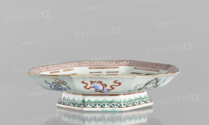 Platter, ceramics, colored porcelain, Qing dynasty, Tongzhi period 1862–1874, Chinese, decorated with the Taoist Taiji sign in the form of two stylized birds intertwined, surrounded by the Eight Immortals and their corresponding trigrams; outer rim decorated with Buddhist symbols. Vessel; height 5.8 cm, diameter 24.5 cm., A shallow, octagonal porcelain dish with a raised foot, displayed against a plain light gray background, showing pale celadon and white glaze with painted decorative motifs in colors including green, blue, red, brown, and gold; the rim has a reddish-brown border pattern, the exterior sides feature a few stylized floral and ribbon-like designs in blue and red with touches of yellow and brown, and the foot is decorated with green wave-like patterns and small pale dots; the dish casts a soft reflection on the glossy surface below.