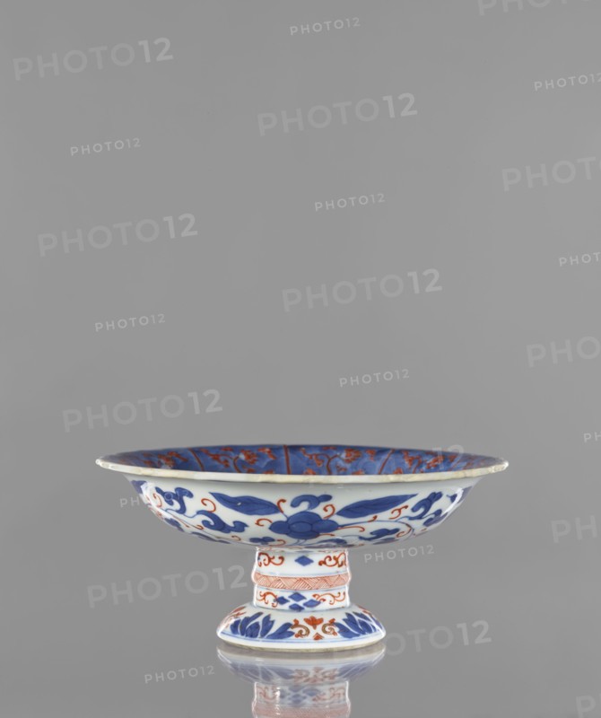 Platter, Narozno Eckersdorf, phoenixes iconography, ceramics, platters, Japanese art, Japanese culture, floral ornaments, colored porcelain, gardens, birds, Edo 1603–1868, early 18th century, Japan, Arita, vessel, height 9.7 cm, diameter 21.7 cm, A shallow pedestal dish sits centered against a plain gray background, showing white ceramic decorated with bold blue floral and foliate motifs and additional orange-red accents; the wide rim flares outward and the interior shows blue panels with orange-red detailing, the exterior of the bowl displays flowing blue leaves and scrolls punctuated by orange-red curls, and the short cylindrical stem and round foot are similarly ornamented with bands of blue leaves and orange-red decorative patterns, with a subtle reflection of the piece visible on the surface below.