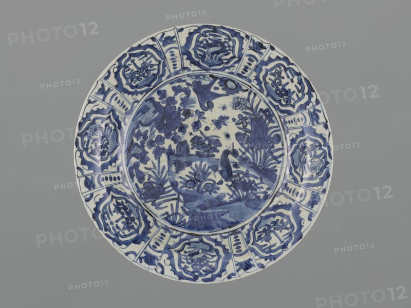 Platter, Safavid dynasty 1501–1736, Persian; ceramics, round deep platter with separate rim on a ring foot. Decoration: in the mirror a pond scene with rocks, three ducks and various aquatic plants and insects; on the collar in eight multi-leafed cartouches cranes and stylized plants; stylized plants on the outer side of the rim. On the reverse a handwritten inscription in Arabic script; within the ring of the foot the words jami'-je Bali handwritten in brown dye, likely referring to an unspecified locality. The asymmetric composition—central rock and a bank jutting into the water with a duck—maintains a circular form without horizontal landscape division. Decoration imitates motifs of blue-and-white Chinese kraak porcelain and alludes to Chinese painting, but Persian ceramic depictions are freer and denser in motifs; plant-animal compositions birds, insects, quadrupeds echo Persian textile and miniature traditions. Nature was a frequent Safavid subject; the first landscape composition...