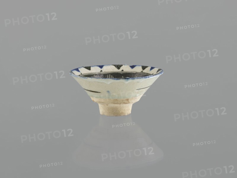 Bowl, Islamic art, Arabic culture, Ayyubid dynasty 1171–1341, ceramics, Arabic ceramics, Middle Eastern ceramics. Bowl on a ring-shaped foot, wide bottom, inverted-cone shape characteristic of Syrian bowls and goblets produced during the Ayyubid period. Likely quartz ceramic mass probably made in Egypt in the 12th century, a feature of Ayyubid wares. Decoration: stylized plant motifs — a lotus flower and a leaf — in the mirror, surrounded by rays of light; linear and radial ornament painted in black; stylized flower in the mirror. Decoration executed in a free brush style popularized by Syrian artists in the 12th–13th centuries; composition shows formal simplification typical of contemporary Middle Eastern ornamentation. M.P., 13th century, Syria; vessel; weight 0.26 kg; height 7.4 cm; diameter 15.9 cm., A small shallow ceramic bowl sits centered on a smooth reflective surface, showing a muted reflection below. The bowl has an off-white to cream base color with a slightly rough unglaze...