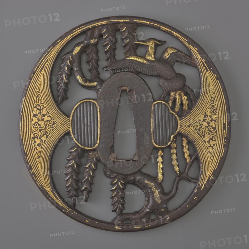 Crossguard tsuba, Japanese, Edo period 1603–1868, early 18th century, weapons — katana crossguard tsuba, maru gata round, kaku mimi rectangular edge, ryôhitsu-ana oval blinded with shakudô. Decorated in positive openwork yô sukashi with a hoo phoenix perched on a willow tree; decoration and edge have gold inlay using the nunome technique. Aizu-Shôami style. Not signed. Dimensions: height 8.4 cm, width 8.2 cm, depth 0.45 cm., A round dark metal object with a central oval opening and an irregular cutout, decorated with areas of gold detailing; a stylized bird with outstretched wings appears near the top right, and a group of hanging, leaf-like forms dangles toward the lower left and center; two rounded, ribbed panels flank the central opening and are outlined in gold; ornate, fan-shaped gold patterns fill the left and right edges of the circle; the surface shows textured, carved branches and organic shapes, with a color palette of dark brown or black metal and bright gold accents against...
