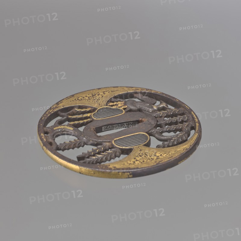Crossguard tsuba, Japanese, Edo period 1603–1868, early 18th century, weapons — sword crossguard tsuba, maru gata round, kaku mimi rectangular edge. Aizu-Shôami style. Tsuba with oval ryôhitsu-ana blinded with shakudô. Decorated in positive openwork yô sukashi with a hoo phoenix perched on a willow tree; decoration and edge embellished with gold inlay using the nunome technique. Not signed. Materials/technique: metal shakudô, gold inlay. Dimensions: height 8.4 cm, width 8.2 cm, depth 0.45 cm. Japan., A circular carved object with an openwork design showing a few curved and linear cutouts, with decorative raised textured areas and floral-like motifs; the object has a dark brown base color with several areas of gold detailing and a few worn spots where the gold appears faded, set against a smooth light gray background.