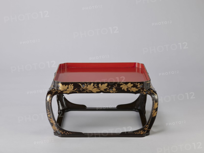 Tray on legs kakeban, noted. Japanese, Edo period early 19th century. One of a set of three tables placed one on top of the other; almost square red tops with high edges and bow legs connected at the bottom by a square frame forming the base. Sides and legs covered with black lacquer and sprinkled with gold using the maki-e technique with flowering cherry plum? and pine branches, depicting plum trees, flowers, trees, pines, twigs, winter. Piece of furniture. Height 25.3 cm; width 47.5 cm; depth 46.0 cm., A low rectangular tray table with a glossy red top set into a black frame, the black legs and apron decorated with gold floral and vine motifs, the legs curving inward to form open rectangular supports, the overall color palette including red, black, and gold against a light gray background.