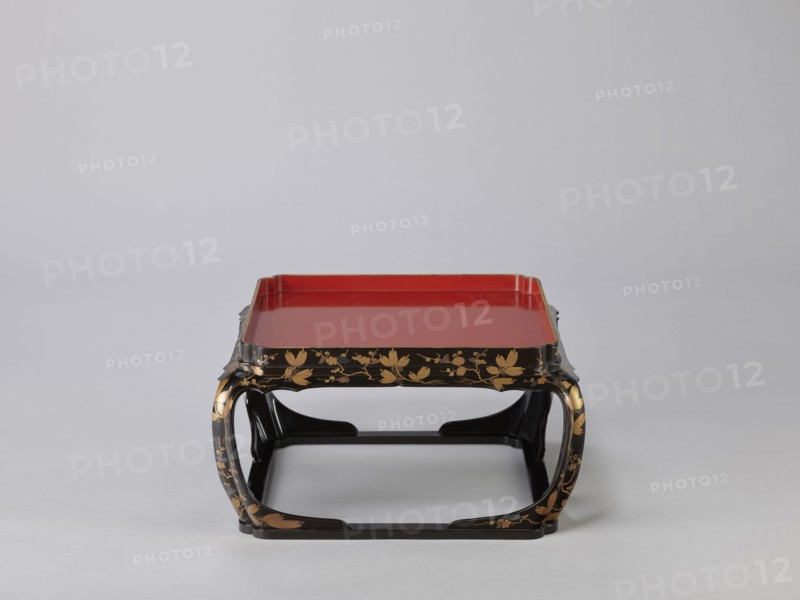 Tray on legs kakeban, Japanese, Edo period early 19th century. One of a set of three tables placed one on top of the other; almost square red tops with high edges and bow legs connected at the bottom by a square frame forming the base. Sides and legs covered with black lacquer and decorated with gold powder using the maki-e technique in a motif of blooming plum and pine branches, depicting plum trees, flowers, trees, pines, twigs, winter. Piece of furniture. Height 22.0 cm; width 41.5 cm; depth 40.0 cm., A decorative low table with a red flat top set into a black frame that curves into arched legs, the black surfaces embellished with gold floral and leaf patterns; the tabletop has raised edges and the overall scene shows a neutral light gray background and floor.