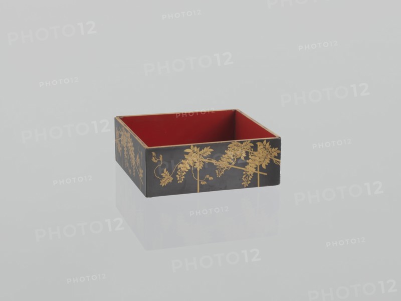 Portable picnic set sagejûbako, Rectangular cabinet without side walls on a stand with two stacked food containers set incomplete; originally four; beside them on the stand are two tin sake bottles. Top tabletop has a handle, one small drawer, and a shelf for trays missing. Entire exterior covered with black roiro-nuri lacquer; interiors of food containers with vermilion lacquer. Surface ornament executed in hiramaki-e and nashiji techniques, depicting blooming wisteria, camellias, plum trees, wolfberries, mon rosettes/coins, and leaves. Such portable eating sets were variously called in Japan kuchi?, sagej?, hanami bent? and were popular from the Momoyama period mid-16th–early 17th c. and in the 18th–19th centuries. 19th century Edo–Meiji periods, Japan. Dimensions: height 31.3 cm; width 30.5 cm; depth 17.8 cm; weight 2.29 kg., A rectangular open box with a red interior and a black exterior decorated with gold floral patterns sits on a reflective pale surface, showing a faint mirrored...