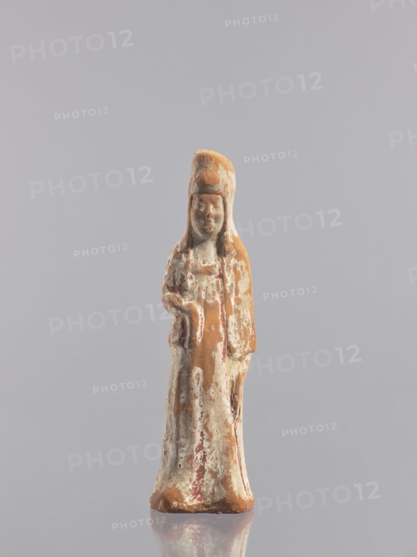 Standing woman figure - tomb figurine, Chinese, ceramics, female figurine, Tang dynasty 618–907, early 10th century, China, sculpture, height 19.3 cm, A small weathered figurine stands upright against a plain light gray background, showing a long robe and a tall rounded headdress; the surface of the figure is a mix of earthy brown, off-white, and muted reddish tones with areas of worn texture and flaking, and the base reflects faintly on the smooth surface beneath it.