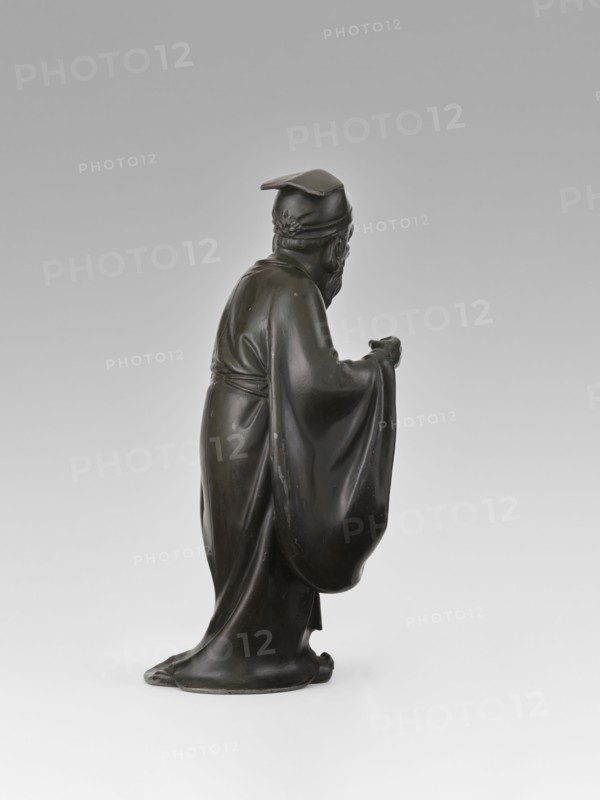 Sage, unknown sculptor, figurine, Chinese art, Chinese culture, unknown provenance; subject: sage man, scholar wearing a long flowing robe and a cap on his head; Qing dynasty 1644–1911, 19th century; material: sculpture; height 28.0 cm., A dark bronze statue of a robed figure shown in profile, standing with a long flowing garment that pools at the base and drapes over the arms; the figure wears a cap with a flat top and a decorative band at the back, has a beard and mustache, and one hand is extended forward while the other is held closer to the body; the surface shows smooth highlights and subtle wear, and the background is a light neutral gradient of gray and off-white.