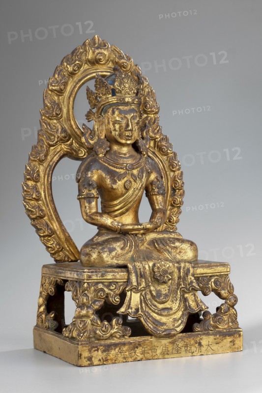 Amitayus, Buddha of Infinite Life, shown seated in a meditative pose with hands folded in a meditative gesture. Qing dynasty 1644–1911, dated 1770. Sino-Tibetan style; Himalayan/Chinese/Tibetan Buddhist art. Sculpture, height 22.0 cm, width 11.5 cm, depth 8.9 cm. Hair styled in a chignon with a high bun tied on top of the head and painted blue; head adorned with a five-leaf crown inlaid with semi-precious stones set on a headband mounted on the underside. Ushnisha present; lotus throne and halo indicated. The back is painted orange. Missing elements include hair flowing to the shoulders, the sash covering the shoulders, stones, and earrings probably large, flower-shaped, falling on the shoulders. The bottom of the sculpture is closed with a wooden plaque secured with metal pins and mortar; the plaque bears a drawing of a quadruple vajra., inscriptions present., A gilded statue of a seated figure on a decorative platform, showing a figure with a serene face, closed eyes, and detailed ha...