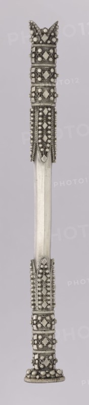 Hookah mouthpiece; maker, Islamic art; Arabic; smoking accessory hookah/pipe; geometric ornament and pearling; early 20th century; likely Yemen or Arabia Petraea origin; length 18.1 cm; diameter 2.1 cm., A vertical metallic object with an elongated central shaft in a pale silver color and decorative end sections in a darker silver tone; the top end features a crown-like cluster of rounded beads and faceted diamond-shaped elements, surrounded by coiled wire and rows of small spherical beads, with textured raised bands and looped metal details; the lower end mirrors the top with similar patterned bands of coiled wire, clusters of small round beads, and diamond-shaped facets culminating in a rounded base with a ring of larger beads; the central shaft is smooth and reflective, showing subtle linear facets and a uniform pale silver finish against a light muted background in a very light grayish-lavender tone.