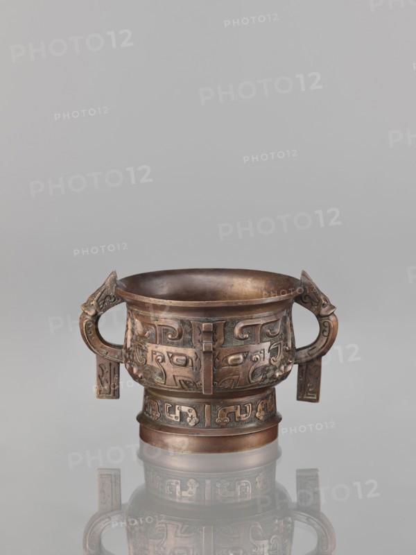 Gui-type incense burner, bronze, Chinese, Qing dynasty 1644–1911, 19th century. Gui vessel with taotie monster and fantastical beast motifs, ritual/ceremonial vessel, height 10.0 cm, width 11.5 cm. Creator, ., A bronze ritual vessel with a rounded bowl on a short cylindrical foot standing on a reflective surface, showing a patina of brown and dark brown with areas of greenish oxidation; the vessel has a pair of high loop handles with stylized animal heads at the terminals and rectangular pendant plaques hanging from the handles; the body and foot are decorated with repeating raised abstract motifs and scroll-like patterns in low relief; the interior appears darker brown and smooth, while the exterior shows subtle variations of brown, dark brown, and hints of green, and the piece casts a muted reflection on the pale gray background.