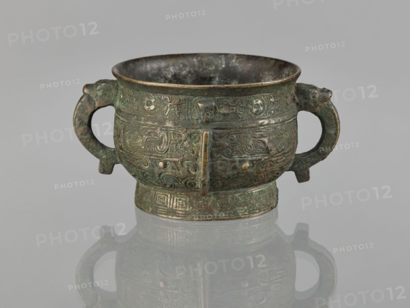 Gui incense burner, Chinese, ceremonial ritual vessel gui, 19th century Qing 1644–1911, taotie and meander leiwen ornamentation, material unspecified, height 6.5 cm, width 12.8 cm, diameter 8.9 cm., An aged metal vessel with a rounded bowl and a short flared rim sits centered on a reflective surface, showing a mirrored image beneath; the vessel has two looped handles on opposite sides and a short cylindrical base, all covered in intricate raised decorative patterns and motifs in a worn, textured finish. The visible colors include greenish patina, dark brown, muted bronze, and hints of black, with areas of lighter metallic sheen where the surface is more exposed. The interior appears darker and mottled, the surface shows irregular corrosion and verdigris, and the reflections on the base echo the vessel’s shape and colors.