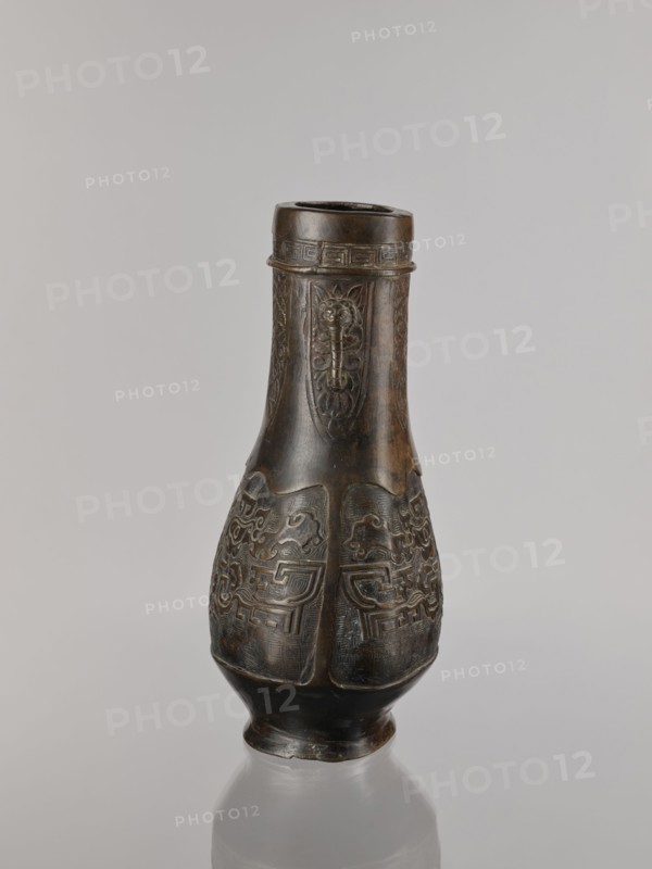 Hu-type vessel, bronzes, Chinese art, Chinese culture, Qing 1644–1911, ceremonial/ritual vessel, taotie iconography, meander ornament, Qing style, 19th century, China, vessel, height 28.0 cm, width 19.0 cm, A carved vase-like object stands upright on a reflective surface, showing a dark brown to nearly black patina with lighter brown highlights on raised areas; the object tapers from a wider rounded midsection to a narrower neck and flared rim, and the body features repeated carved panels with stylized animal or abstract motifs and patterned borders, while the neck displays elongated vertical decorative panels and a band of geometric ornament near the rim; the base is slightly flared and the overall background is a soft gray.