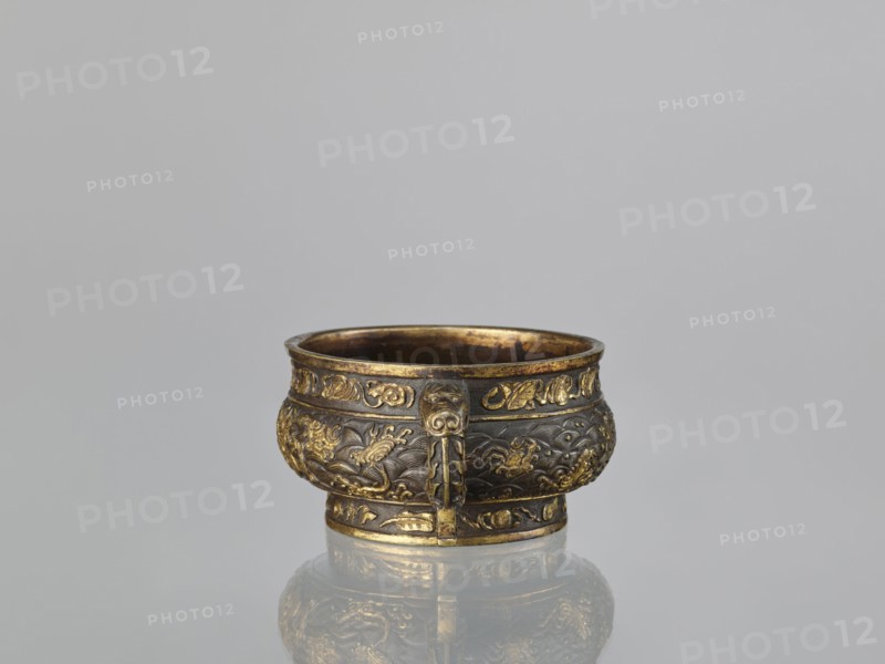 Gui-type incense burner hu, attributed to Hu Wenming fl. ca. 1583–1614. Chinese, 19th century, Qing dynasty. Bronze ritual vessel used as an incense burner. Decorated with dragons, phoenixes, unicorns qilin, plant ornament, sea waves, and hundred antiquities baigu motifs. Height 7 cm; width 18.6 cm; diameter 13.1 cm., An ornate metal bowl sits centered against a plain light gray background, its surface showing a mix of dark bronze and bright gold tones; the bowl has a rounded body with a slightly flared rim and a short base, and it features raised decorative bands and embossed motifs around its sides, including flowing wave-like patterns and stylized organic shapes; a vertical decorative strip with a face-like element projects slightly from the front, and the interior and rim show a warm golden sheen; the bowl rests on a reflective surface that mirrors its colors and shapes.
