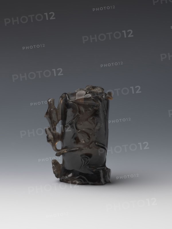 Vase on base, carved ornaments, flowers, birds decorative motifs, stylized rocks; wooden base, oval on three legs; Qing style Qing dynasty, 1644–1911; vessel; 18th–19th century China; height 11.8 cm, diameter 6.3 cm., A dark, glossy carved object resembling a short vertical cylinder with irregular contours sits on a smooth light gray surface against a gradient gray background; the object has organic carved motifs with flowing curved lines and raised rounded forms, and features protruding elements on one side that create openings and negative space, with visible color variations in deep brown to near-black and small areas of lighter brown translucence.