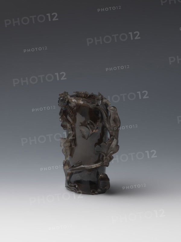 Vase on base, carved ornaments; wooden base, oval on three legs. Carved decoration: stylized rocks, flowers, birds. Chinese art; Qing style Qing dynasty, 1644–1911. 18th–19th century, China. Vessel. Height 11.8 cm; diameter 6.3 cm., The image shows a carved dark brown translucent object with an irregular organic shape, featuring sculpted leaf-like and vine-like forms wrapping around its sides and a small rounded protrusion near the base; the object sits on a smooth surface and is lit against a gradient background that transitions from dark gray at the top to light gray at the bottom, with visible colors including dark brown, gray, black, and hints of lighter brown in the object's surface reflections.