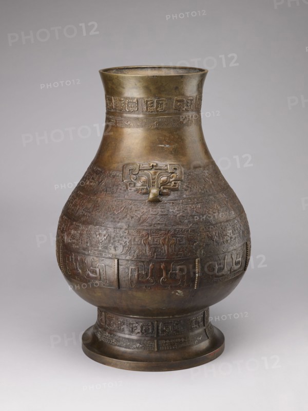 Hu-type vase, Chinese, Qing 1644–1911, 18th–19th century. A hu-type ritual bronze vessel with an ovoid-shaped belly, cylindrical neck with a straight rim, and a high conical foot. Semicircular clubs are attached to the collar. Rich relief decoration on the belly emphasized with engraving; classic decorative motifs include taotie masks, mythical kui animals, stylized birds, dragons, Chinese symbols, and geometric patterns. Ritual vessel, height 59.5 cm, width 40.0 cm, weight 19.0 kg., A large rounded metal vessel with a flared neck and a pedestal base sits against a neutral gray background; the vessel displays a dark bronze and brown patina with areas of lighter gold-toned metal, and its surface is covered in intricately carved or cast decorative bands and raised motifs including a stylized animal or mask-like form on the upper body, with repeating geometric and curvilinear patterns encircling the midsection and base, and subtle variations of darker brown, reddish-brown, greenish-brown,...