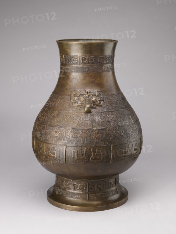 Hu-type vase, Chinese art, Chinese culture; subject: birds, dragons, taotie, Chinese symbols; function: ceremonial/ritual vessel bronze hu-type; description: ovoid-shaped belly, cylindrical neck with straight rim, high distinct conical foot; semicircular clubs attached to the collar; rich relief decoration on the belly emphasized with engraving; classic decorative motifs: taotie masks, mythical kui animals, stylized birds, geometric patterns; period: Qing 1644–1911, 18th–19th century, China; dimensions: height 59.5 cm, width 40.0 cm, weight 19.0 kg., A large, rounded metal vessel with a narrow flared neck and a broad foot stands against a plain light gray background; the vessel shows an overall brown and bronze patina with areas of darker and lighter bronze tones, and a few subtle greenish and golden highlights. The body of the vessel is covered in repeated raised decorative bands and intricate carved or cast geometric and zoomorphic patterns encircling the midsection and neck, and a s...