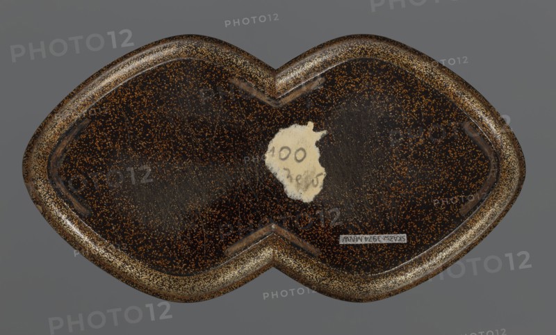 Tray, lacquerware, Japanese art; tray with asymmetrical decoration of an old plum tree trunk with young shoots and birds kachôga decorative motif; Edo period, 1st half of the 19th century; Japan; vessel; height 2.6 cm, width 17.8 cm, depth 9.5 cm., A close-up view of a heart-shaped object with a dark brown surface speckled with gold or tan flecks, set against a flat gray background; the object has a glossy finish with visible surface scratches and a worn spot revealing a lighter beige area, and its raised rim shows denser gold flecking and a slightly darker brown edge.