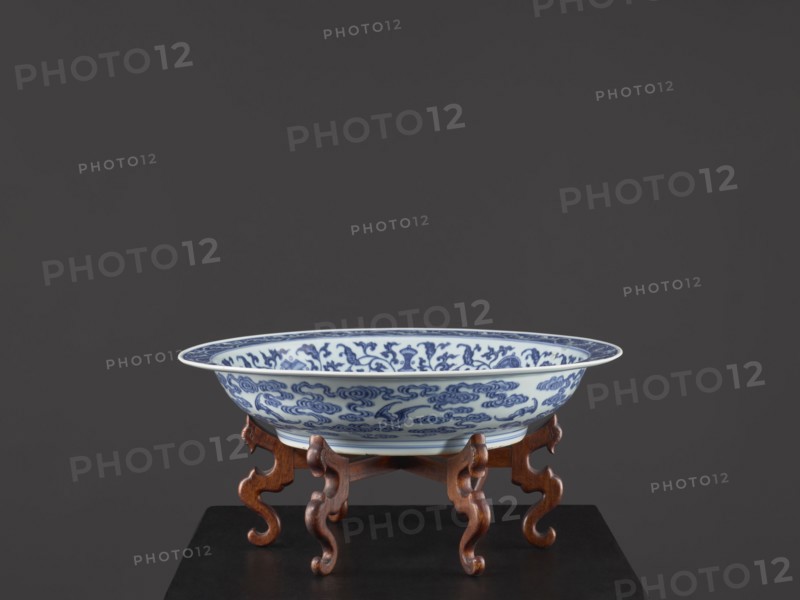 Platter on stand, Blue-and-white porcelain platter on a low foot with a wide rim, sitting on a carved wooden stand. Decorated with plant tendrils, bats, and Buddhist emblems. Center of the mirror features a rosette filled with an archaized swastika ornament; outer wall shows bats among clouds. Qing dynasty, Yongzheng period 1723–1735, China. Vessel; height 21 cm, diameter 45.5 cm., A shallow porcelain bowl with blue decorative patterns sits on a carved wooden stand; the bowl is primarily white with intricate blue designs on its interior and exterior, the stand is a warm brown wood with curving legs, and the background is a dark gray.