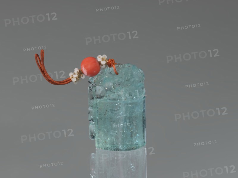 Seal stamp pendant, Chinese, Qing dynasty 1644–1911. Subject: lion iconography. Materials: aquamarine stone, carved and polished in the upper part; strung with river pearls and coral. Function: seal stamp. Date: 18th–19th century. Dimensions: height 4.1 cm, width 1.8 cm, depth 2.7 cm., A translucent bluish-green cylindrical stone with an irregular carved top sits upright on a reflective surface, topped by a loop of reddish-brown cord threaded through a smooth round reddish bead and accented by small white bead clusters; the background is a smooth gradient of gray.
