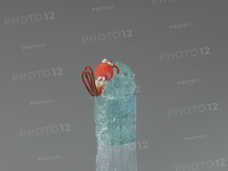 Seal stamp pendant, Chinese, Qing dynasty 1644–1911; carved stone aquamarine, polished in the upper part; carved lion iconography; drilled for suspension with string featuring river pearls and coral; 18th–19th century; seal stamp; dimensions: height 4.1 cm, width 1.8 cm, depth 2.7 cm., A small translucent carved object in a pale aqua color sits upright on a reflective surface, topped by a few decorative elements including a round bright coral-red bead, a few tiny white flower-shaped beads, and a looped cord in a rusty orange-brown hue; the background is a smooth gradient of gray.