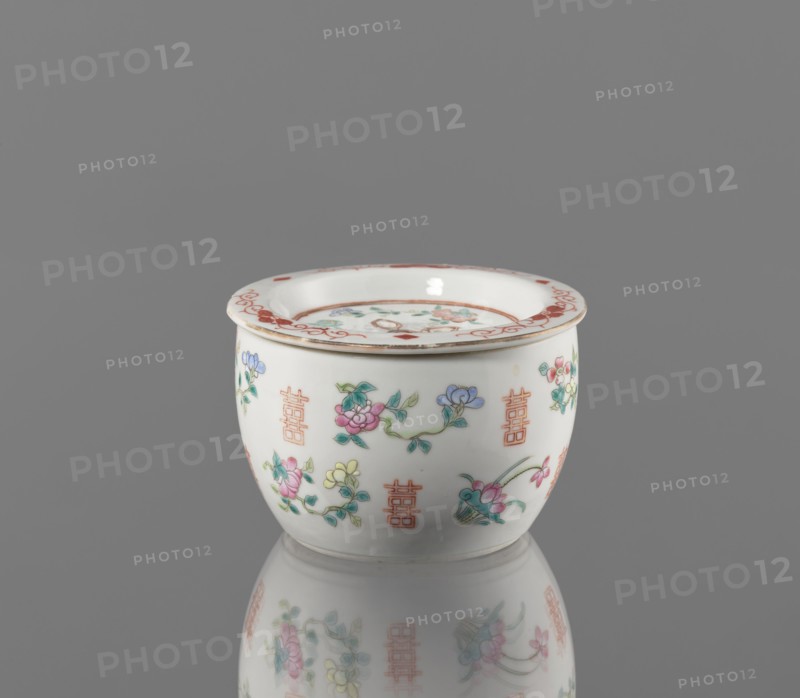 Hand warmer, colored porcelain, Qing dynasty Tongzhi period, 1862–1874, China. Ceramic vessel with bulging body and flat recessed lid pierced with five holes; decorated with floral motifs—peonies, lotuses—and peaches as longevity shou iconography. Height 7.3 cm; diameter 11.2 cm., A small white ceramic bowl with a flared rim sits on a reflective gray surface, showing a soft mirrored reflection beneath it; the bowl is decorated with delicate floral motifs in pink, blue, green, and yellow, accented with fine reddish-orange decorative marks along the rim and repeated vertical ornamental symbols around the body, and there are touches of gold along the rim.