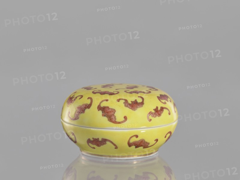 Round box with a convex lid a cinnabar vessel for seals, bats iconography, ceramics, colored porcelain, Qing dynasty, Tongzhi 1862–1874, Chinese, lid, seals, vessel; height 4 cm, diameter 7 cm., A small rounded box with a lid sits on a reflective surface; the box is yellow with a repeating decorative motif in reddish-brown, and the background is a uniform light gray.