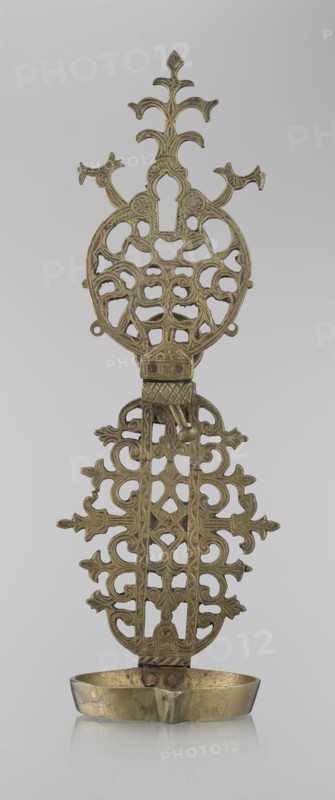 Oil lamp, Persian/Islamic art, 19th–20th century, Central Asia or Iran, oil lamp with side decorative shaft, stylized medallion and palmette finial; decorated with birds, geometric motifs and palmettes; height 45.5 cm, diameter 15.5 cm., A vertical metal object with an ornate openwork design and a shallow bowl at the base, shown against a plain light gray background; the metal has a muted gold to brass color with areas of darker patina and slight discoloration; the top portion features symmetrical curving branches and rounded cutouts forming a decorative crest, followed by a central section with repeating open scrollwork and stylized floral or foliate motifs; a small hinge-like element connects the upper and lower panels, and a few small protruding pins or rivets are visible where the bowl meets the main structure; the shallow bowl at the bottom is oval with a gently curved lip and shows subtle surface marks and a slightly uneven sheen.
