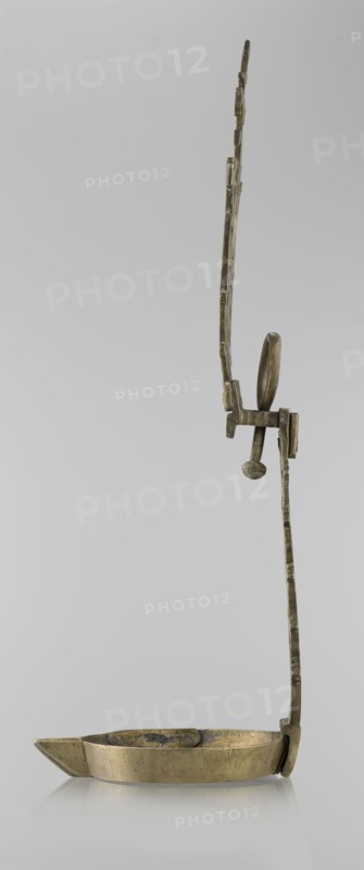 Oil lamp, Persian Islamic art, oil lamp for lighting with birds, geometric motifs, palmettes ornament; lamp with a side decorative shaft, stylized medallion, palmette finial. 19th–20th century, Central Asia, Iran, height 45.5 cm, diameter 15.5 cm., A metallic object with a vertical elongated arm and a shallow round dish at the base, shown against a light gray background; the object appears made of a brass or bronze metal with a dull golden-brown patina and areas of darker brown and greenish tarnish, the vertical arm is thin and irregular with a few small protrusions and a rounded knob near the middle where a horizontal element connects, the shallow dish is circular with a flat rim and a small triangular spout on one side, the metal surface shows scratches and worn spots and some darker patches inside the dish.