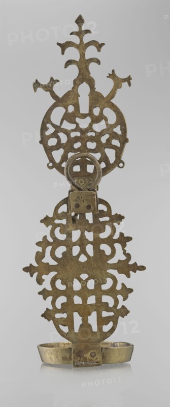 Oil lamp, Persian, 19th–20th century, possibly Central Asia or Iran; ceramic/metal oil lamp with side decorative shaft, stylized medallion and palmette finial; decorated with birds, geometric motifs, and palmette ornament; height 45.5 cm, diameter 15.5 cm., An upright metal object with an ornate openwork design and a hinged ring near the top, featuring a decorative top section with branching shapes and a larger symmetrical middle section of repeating cutout patterns, all showing a mottled metallic gold and brown patina; a small rectangular hinge piece connects to a lower ornamental panel with similar cutout motifs, which sits above a shallow metal tray with a slightly curved rim that shares the same gold and brown coloring; the background is light gray.