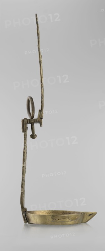 Oil lamp, Islamic art, Persian, oil lamp for lighting decorated with birds, geometric motifs, palmettes; lamp with a side decorative shaft, stylized medallion, palmette finial. 19th–20th century, Central Asia and/or Iran; height 45.5 cm, diameter 15.5 cm., A metallic object with a long thin vertical arm that has an uneven, textured surface and a looped handle near its midsection, attached to a horizontal bracket and a short peg ending in a rounded knob, connected at the base to a shallow, oval dish with a small pointed spout; the object is primarily brass-colored with areas of darker patina and faint greenish and brownish tones, set against a plain light gray background.