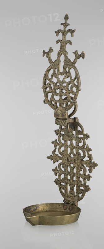 Oil lamp, Islamic art, Persian, oil lamp with side decorative shaft and stylized medallion, palmette finial; decorated with birds, geometric motifs, and palmettes; 19th–20th century, possibly Central Asia or Iran; height 45.5 cm, diameter 15.5 cm., An ornate metal object with an elongated decorative backplate featuring openwork filigree and floral-inspired motifs in metallic gold and aged brass tones, attached to a shallow oval dish at the base that shares the same gold-brass coloration and shows a slightly worn surface and darker patina in places.