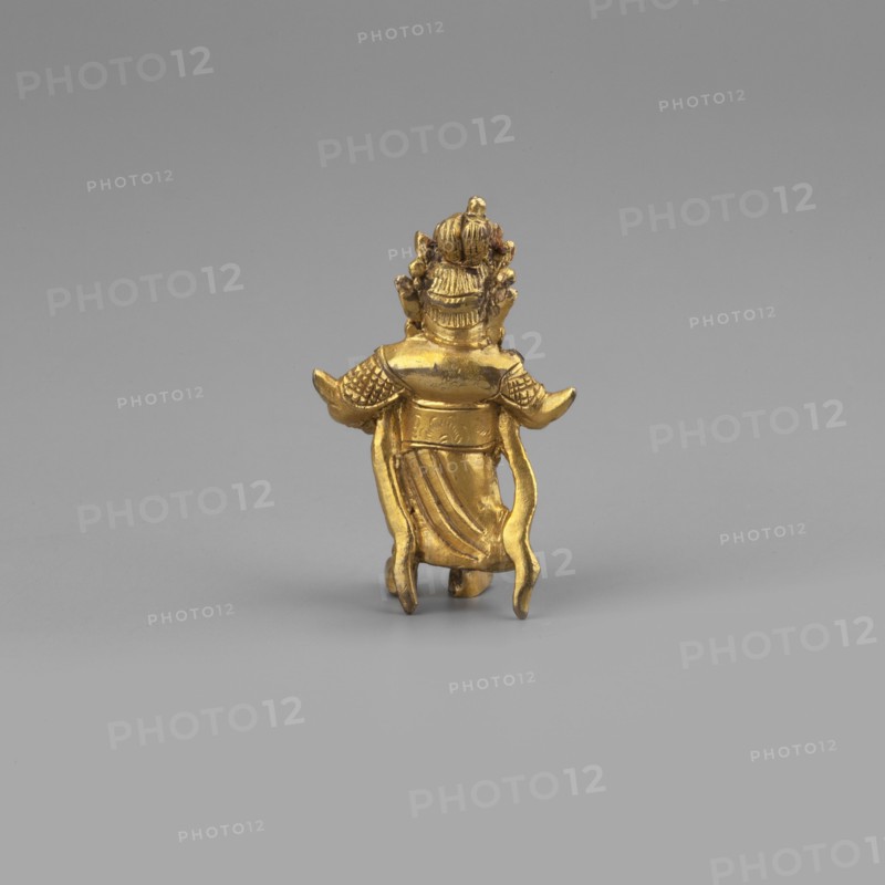 Vaisravana Tib. Namtose, Tib. rnam thos sras, sometimes identified with the yellow Jambhala, an emanation of Buddha Ratnasambhava — guardian of the north. Sculpture figurine, cult object, height 3.4 cm. Himalayan/Tibetan Buddhist art; 2nd half of the 18th–19th century. Maker, provenance: purchase. Possible origin: Tibet or Mongolia., A small gold-colored metal figurine viewed from the back standing against a plain light gray background, showing a figure with an ornate headdress, textured shoulder armor, flowing drapery down the back, and raised arms with curved sleeve-like extensions; the colors visible are gold on the figurine and shades of light gray in the background and surface.