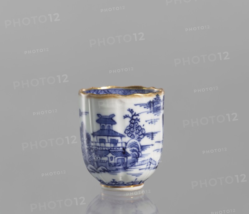 Cup, ceramics, blue-and-white porcelain; Qing dynasty, 19th century Qing, 1644–1912; Chinese; vessel with landscape motifs; part of a 22-piece tea and coffee set including a teapot, 6 cups, 6 saucers, a bowl, a cover, and a plate; height 6.5 cm, width 5.9 cm, diameter 5.9 cm., A small porcelain cup with a scalloped rim edged in gold sits against a plain gray background, decorated in blue and white with a detailed landscape scene including a pagoda-like building, trees, and architectural elements; the interior shows blue patterning and the base also has a gold trim, and the cup casts a soft reflection on the surface below.