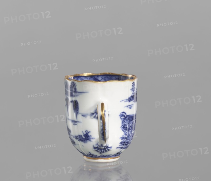 Cup;, Porcelain, blue-and-white; Chinese, Qing dynasty, 19th century. Landscape motifs. Part of a 22-piece tea and coffee service teapot, 6 cups, 6 saucers, a bowl, a cover, and a plate. Vessel. Height 6.5 cm; width/diameter 5.9 cm., A small porcelain cup with a scalloped rim and a single vertical handle sits on a reflective surface against a plain gray background; the cup is primarily white with blue painted landscape motifs and has gold trim along the rim, the handle, and the base, with the interior showing blue decoration and gold edging.