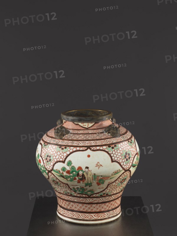 Vase., Colored porcelain with gilded edge. Mascarons on the shoulder; decoration: four reserves with figural representations. Mid-18th century Qing dynasty, 1644–1911, China. Height 40.5 cm; diameter 40 cm. Chinese art; decorative vessel; mascarons; figural scenes., A rounded ceramic jar with a narrow neck sits on a dark surface against a dark gray background; the jar is predominantly white with red, green, and black decorative patterns and painted scenes, featuring red geometric borders and latticework, green floral motifs, and a central painted scene with human figures and plants; the rim and three small sculpted handles at the neck appear dark metallic or bronze in color; overall colors visible include white, red, green, black, and bronze.