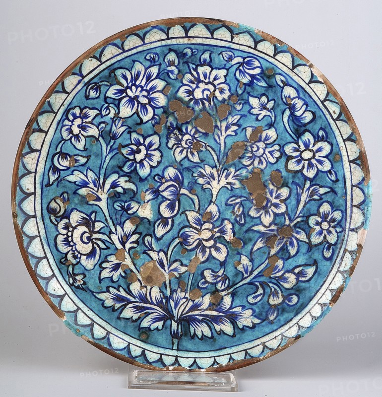 Plate;, Ceramic vessel with height 3 cm and diameter 26.5 cm. 19th century, India/Pakistan . Decoration: composition of a stylized flower bush and crenellated ornament at the edge; floral motifs; plant ornament. Islamic/Indian art., A round ceramic plate with a blue background displaying a floral pattern in darker blue and white tones, with outlines and details in a deep blue or black. The central area shows a cluster of stylized flowers and leaves radiating outward, with petals and foliage rendered in white and blue shading. The rim features a decorative border of scallop-like shapes in pale blue and white with dark blue outlines, and there are several areas of brown spots and worn patches across the surface revealing the underlying material. The plate rests on a clear display stand against a light neutral background.