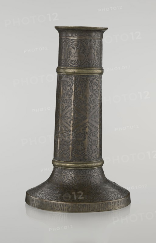 Candlestick mash'al, Islamic art, Persian, Safavid dynasty 1501–1736. Metal candlestick in the form of a column standing upon a low, widening base, its three sections separated by hachured rings. The 10-faceted shaft is decorated with stylized plant ornament and a zigzag; just under the upper edge is a band of inscriptions in nast?aliq script; beneath this are hunting scenes with riders and horses; the background filled with a dense tendril pattern. The base is decorated with representations of huntsmen; an inscription in Armenian characters appears on the base edge, serving a decorative function. 17th–18th century, Iran. Height 28.0 cm; diameter 17.3 cm; weight 0.71 kg., An upright cylindrical metal object with a flared base, shown against a plain light gray background; the object is predominantly dark bronze with areas of muted gold detailing, and its surface is covered in intricate carved or etched repeating patterns and decorative motifs. The midsection and upper rim have raised ba...