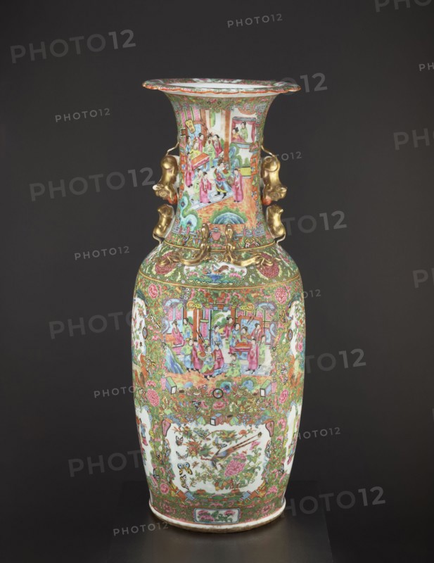 Vase, Chinese, 19th century, colored porcelain vessel with figurative court scenes, butterflies, plant and floral ornamentation; height 93.0 cm, diameter 34.0 cm., A tall decorative porcelain vase stands against a dark background, featuring ornate painted scenes and floral motifs in a palette of pink, green, gold, red, blue, white, and touches of black. The vase has a flared rim and a long neck with painted narrative panels showing groups of people in colorful robes, and the body displays framed panels with birds and floral arrangements surrounded by dense scrolling foliage and blossoms. Gilded sculptural handles shaped like stylized animal heads are attached at the neck, and additional raised gold ornamental elements accent the shoulder area. The lower section repeats floral garlands and intricate decorative borders, and the overall surface is richly detailed with many small motifs and repeated patterns.