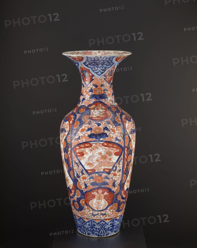 Vase, Japanese, Meiji period ca. 1870–1880, Arita. Colored porcelain Imari, decorated with plant ornaments and floral motifs—peonies, chrysanthemums, pine trees, momiji—and bird images including spotted pheasants and a hoo phoenix; komainu iconography and kachôga themes. Vessel, height 92.7 cm, diameter 37.5 cm, weight 22.5 kg., A tall decorative vase stands against a dark background, showing a glossy surface with intricate painted patterns in blue, red, white, and touches of gold or beige; the vase has a flared rim and a long neck leading to a wider body, with floral motifs, stylized leaves, and rounded medallion shapes arranged symmetrically around its form, alternating panels of dense blue ornamental designs and red floral scenes, and a band of blue patterned decoration near the base.