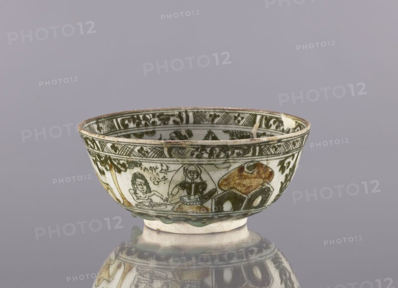Bowl, late 19th century 1893, Iran. Ceramic vessel, height 8.5 cm, diameter 19 cm. Persian/Qajar dynasty. Decoration on the rim: Rostam, with a dragon and Majnun i Lejla and the date 1893. Figural scenes, dragons, birds, literary motifs; utility tableware bowl. Cultural context: Persian/Islamic art., A shallow ceramic bowl with painted decoration sits on a reflective surface, showing a group of human figures and abstract shapes around its exterior; the bowl is primarily white with painted areas in dark green and brown, with touches of yellowish-brown and black, and a rim that shows some wear.