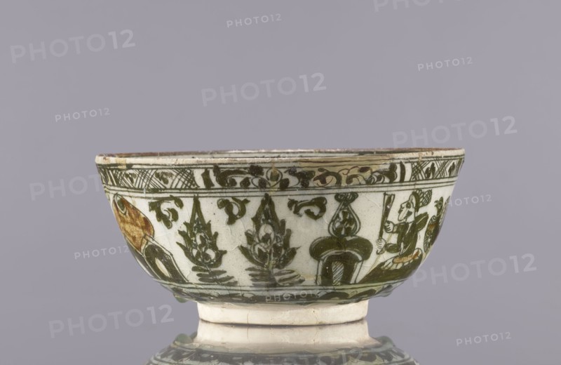 Bowl, late 19th century, Iran. Ceramic vessel, height 8.5 cm, diameter 19 cm. Decoration on the rim: Rostam, with a dragon and Majnun i Lejla and the date 1893. Persian, Qajar dynasty 1785–1925. Figural scenes, literary motifs; imagery includes dragons and birds. Function: tableware/utility ceramic., A shallow ceramic bowl with a flared rim and a short foot rests against a plain mauve-gray background; the bowl's surface is primarily off-white with green painted decorative motifs including stylized plant forms and scrolling patterns, and there are areas of brownish-orange pigment on parts of the decoration; the rim shows a narrow band of darker green ornament and the lower body repeats green motifs above a green horizontal border, while the foot is off-white with some signs of wear.