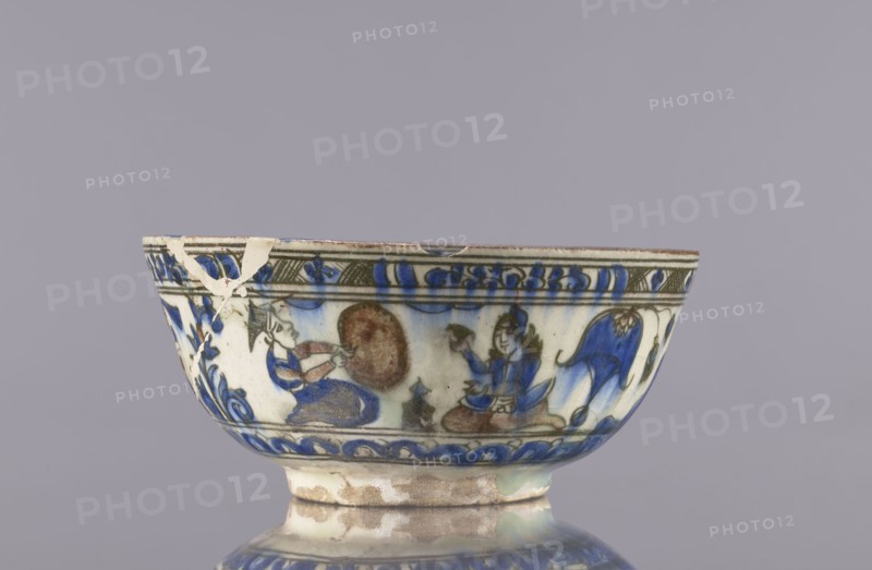 Bowl, Abdullazad Mirza Baba fl. ante 1939, Islamic art, Persian, Qajar dynasty 1785–1925, ceramics decorated with gazelles, figural scenes, lions, female figures; rim decoration: figure with nargile, other figures, lion, palmettes, architecture, inscriptions. 2nd half of the 19th century, Iran. Vessel; height 9 cm, diameter 20 cm., A ceramic bowl with painted decoration sits against a plain light gray background; the bowl shows a band of painted figures and motifs in shades of blue, brown, green, and off-white, with visible glazing and areas of wear and small chips along the rim and body; a few humanlike figures are depicted in blue and brown tones seated on patterned surfaces, accompanied by decorative floral or foliate elements in blue and green; the lower section and foot of the bowl show a mottled off-white and brown surface with glaze variations and some roughened spots.