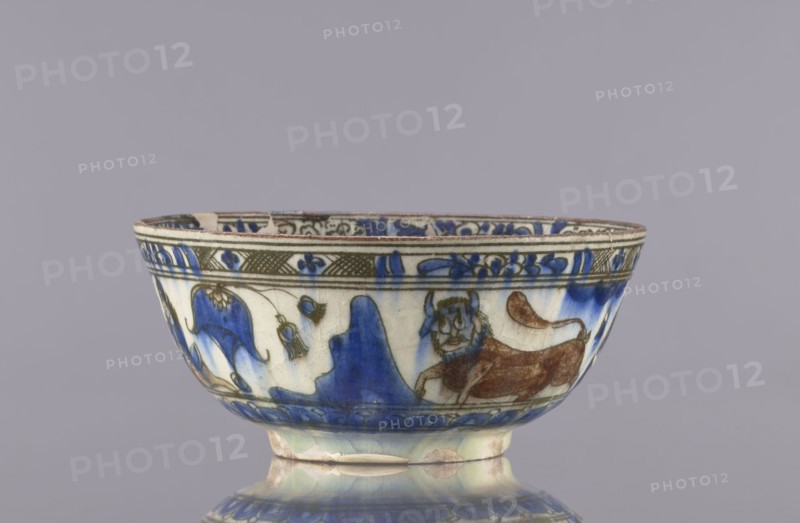 Bowl, Qajar dynasty 1785–1925, 2nd half of the 19th century, Iran; ceramics; vessel, height 9 cm, diameter 20 cm. Decoration: gazelles, figural scenes, lions, female figures; decoration on rims: figure with nargile, other figures, lion, palmettes, architecture; inscriptions. Culture: Persian Islamic art., A shallow ceramic bowl sits on a reflective surface, showing a rounded foot and slightly flared rim; the bowl is decorated with painted motifs including a stylized animal figure and flowing organic shapes. The visible colors include blue, brown, green, white, and hints of beige. The rim and upper band feature patterned ornamentation, while the main body shows a mix of blue washes and brown-painted details forming the animal and surrounding shapes. The glaze has a glossy finish with subtle crackle and areas of wear or discoloration near the base.