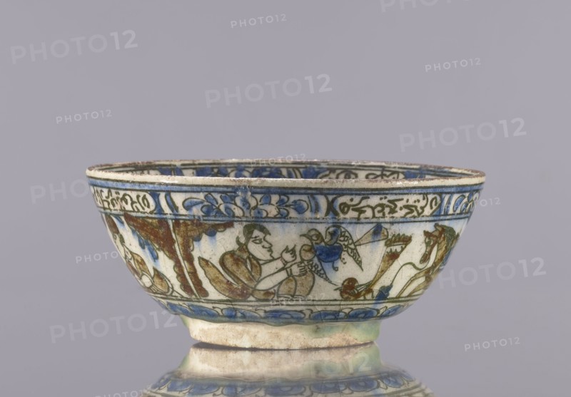 Bowl, Abdullazad Mirza Baba fl. ante 1939, Qajar dynasty 1785–1925, 2nd half of the 19th century, Iran. Glazed ceramic vessel tableware, height 7.5 cm, diameter 17.4 cm. Decoration: knightly and figurative scenes with horses and falcons; rims decorated with figures, architecture, flowers, animals, palmettes, and inscriptions. Persian/Islamic art; ceramics., A shallow ceramic bowl with a rounded foot, shown against a plain light gray background, featuring painted decorative bands and figural scenes around its exterior in colors including blue, green, brown, and cream; the rim and upper band display blue floral and scroll motifs, while the main body shows a group of stylized human figures and animals rendered in brown and green outlines with blue accents, and the lower portion has a repeated blue petal-like border above the foot.